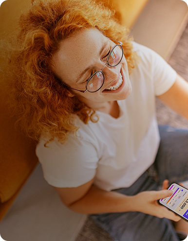 Overhead view of a smiling woman sitting on the floor, holding a smartphone open to the Happen Bank app.